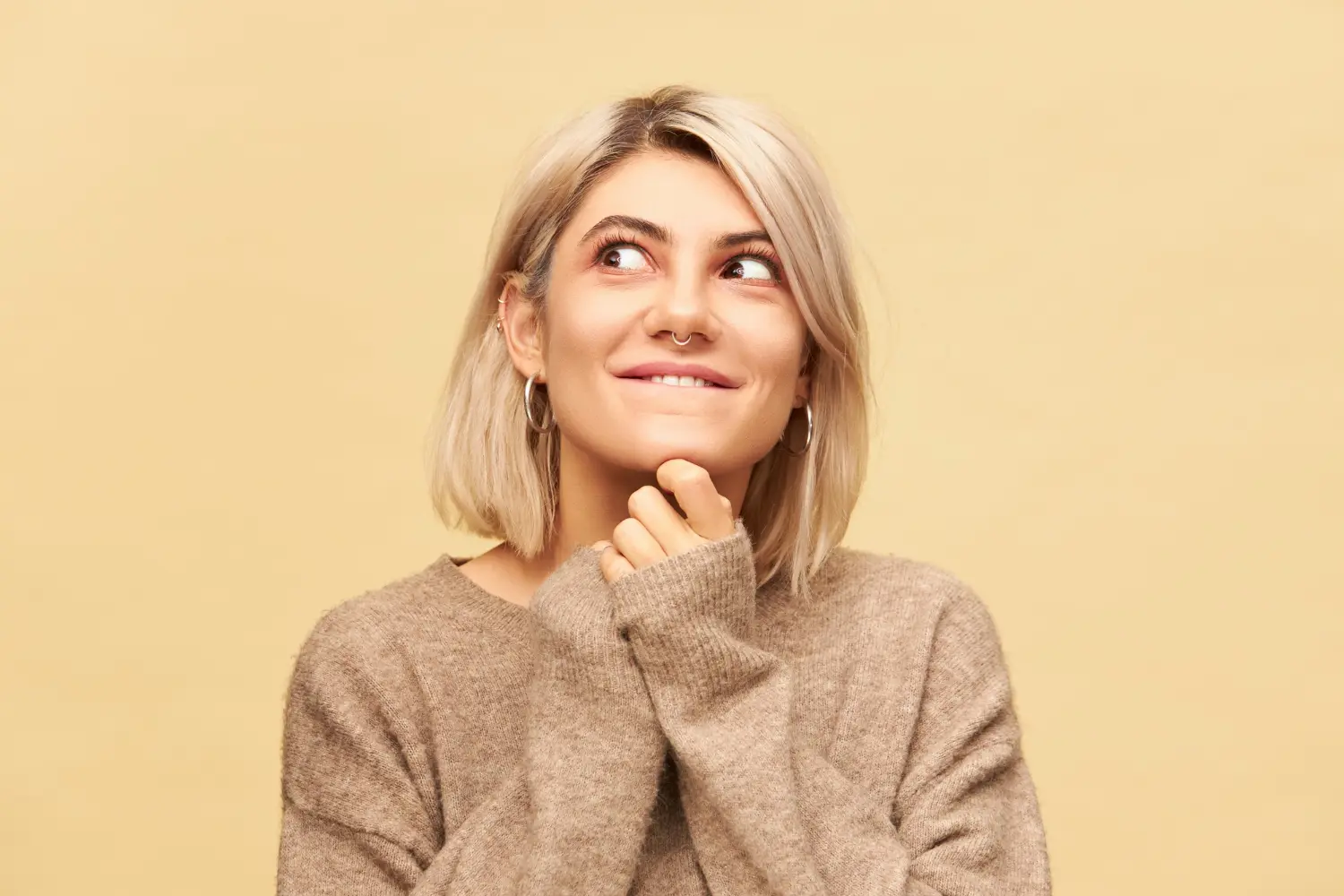 Close up image charming young blonde female wearing nose ring bob haircut holding hands her chin looking away with playful mysterious smile playing pranks doing mischief