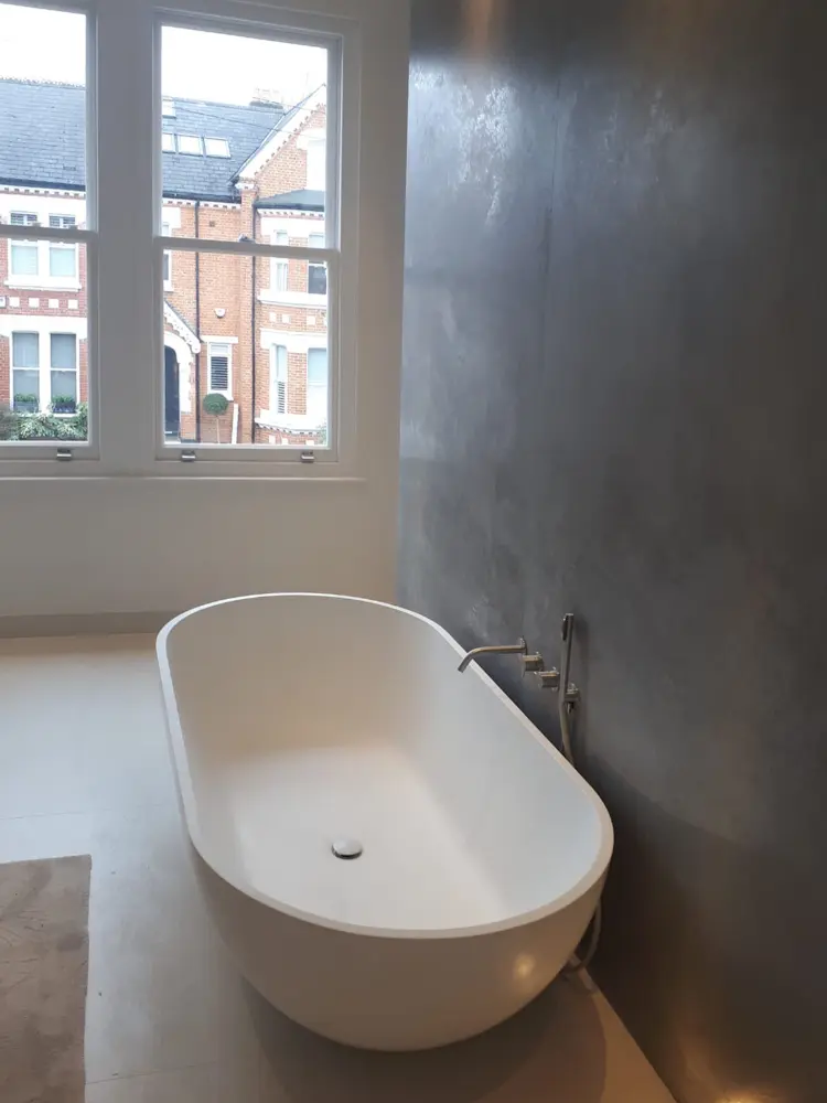 Nowoczesna łazienka z białą, wolnostojącą wanną na ciemnej ścianie i dużym oknem z widokiem na domy na zewnątrz.